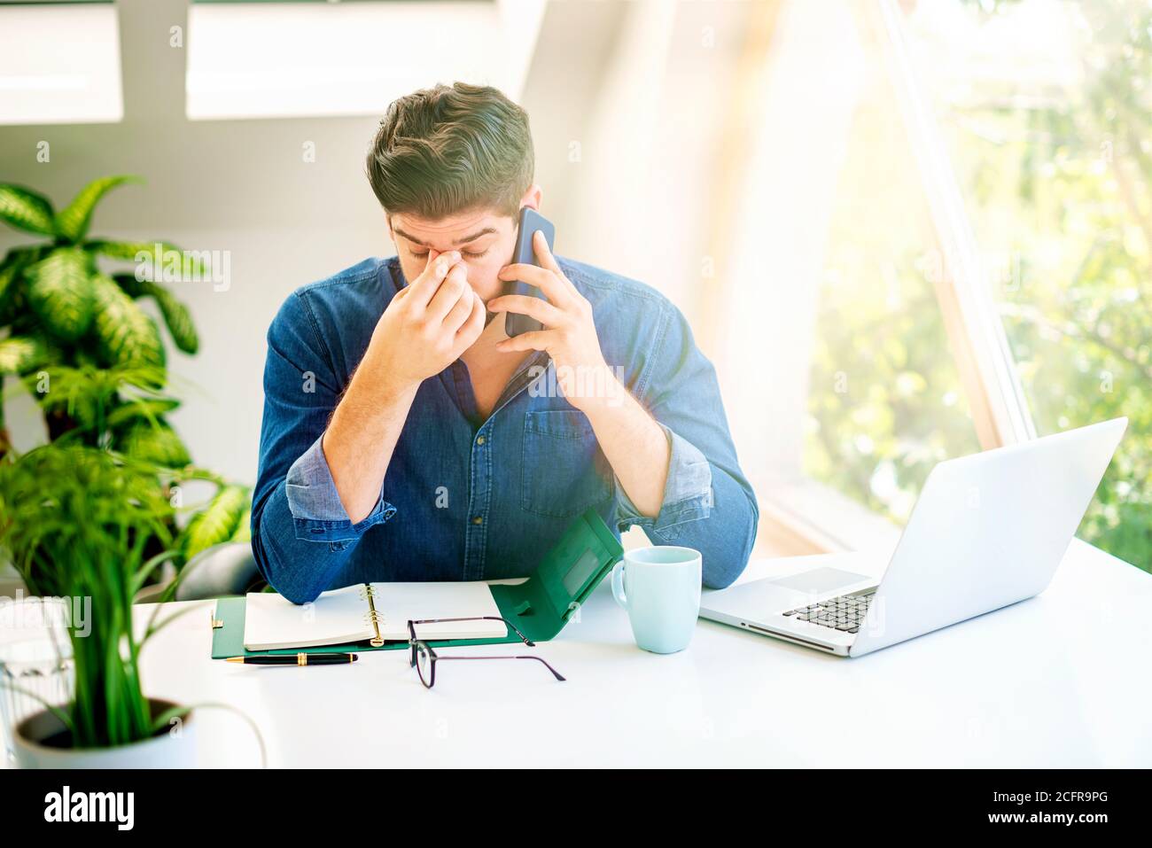 Shot of businessman with hands on forehead sitting behind his laptop and suffering from strong head pain while having a business call. Stock Photo
