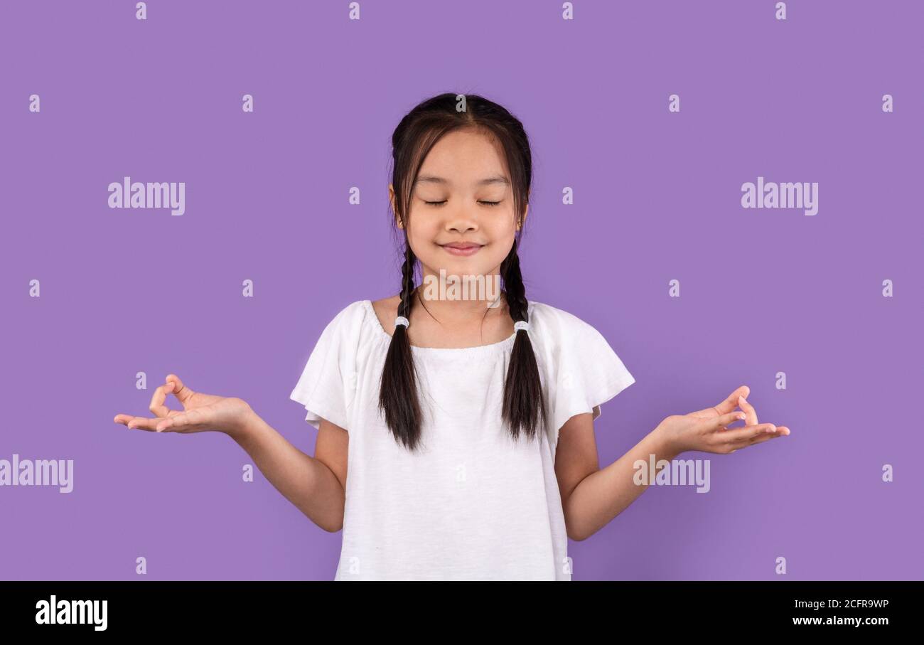 Calm Asian Kid Girl Meditating Standing Over Purple Background Stock Photo