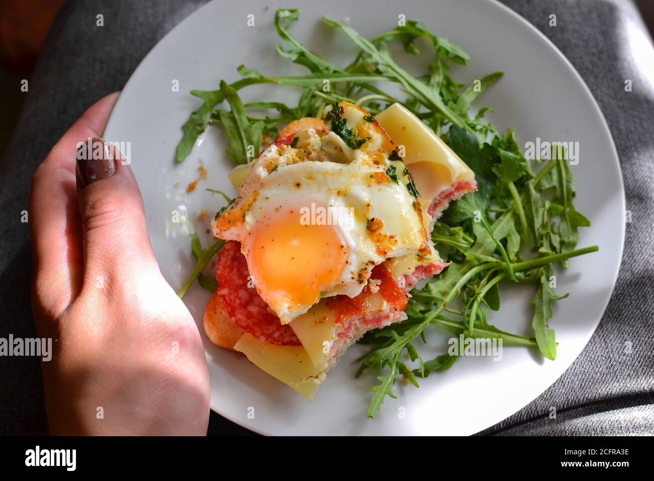 Tasty breakfast. Toast with egg, cheese and sausage. Sandwich in a white plate with arugula. View from above. Light background. Closeup. Woman holding Stock Photo