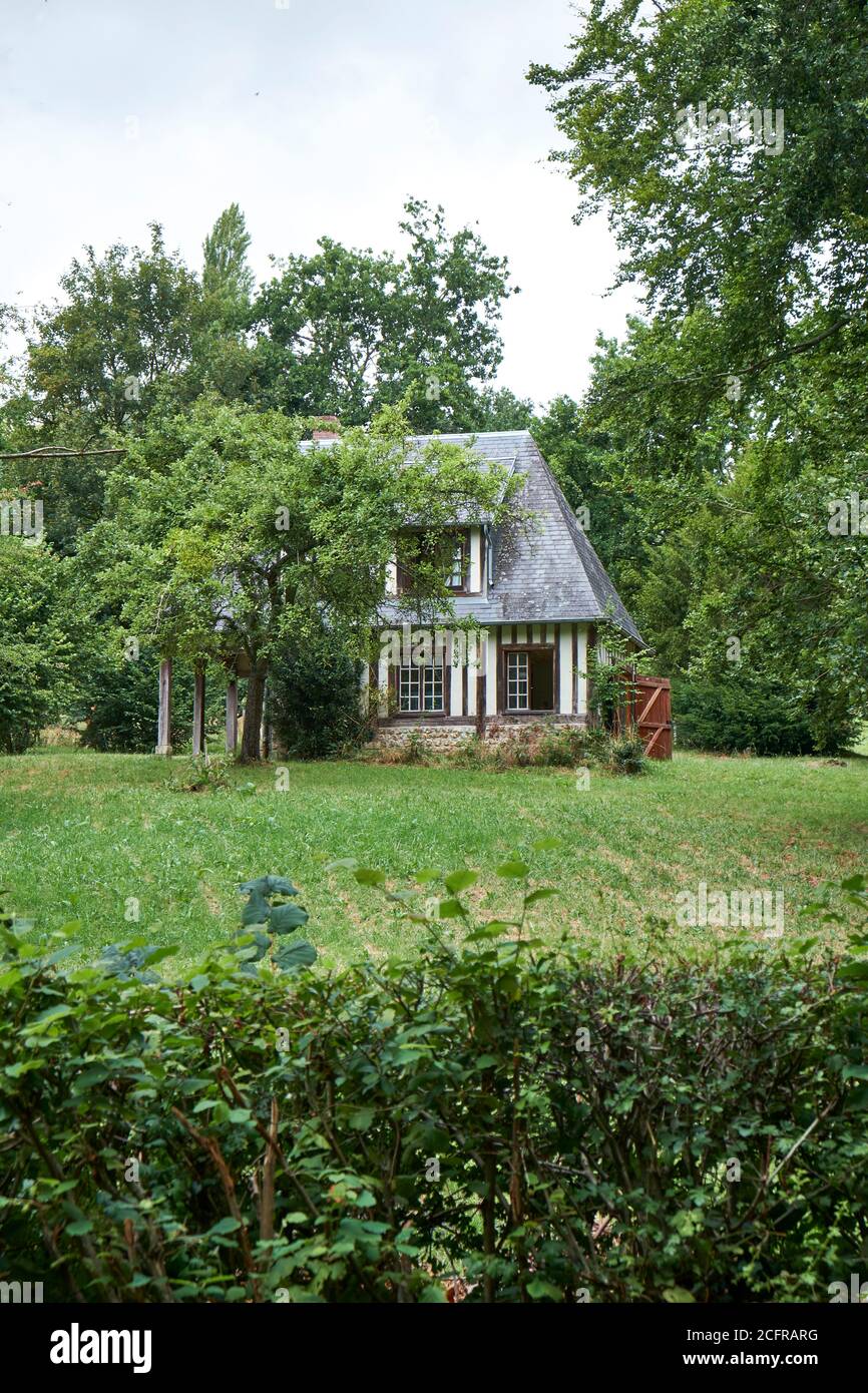 Small detached half-timbered Norman house with slate tiled roof surrounded by trees with garden and hedge in foreground Stock Photo