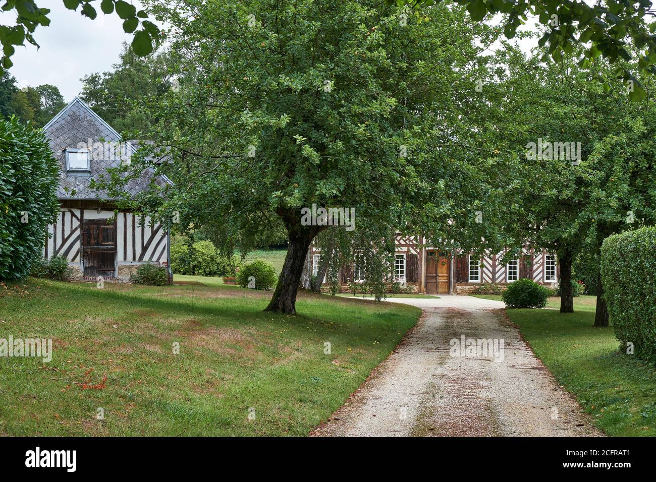 path and driveway flanked by apple trees leading to a half-timbered detached Norman property with freestanding outhouse. Stock Photo