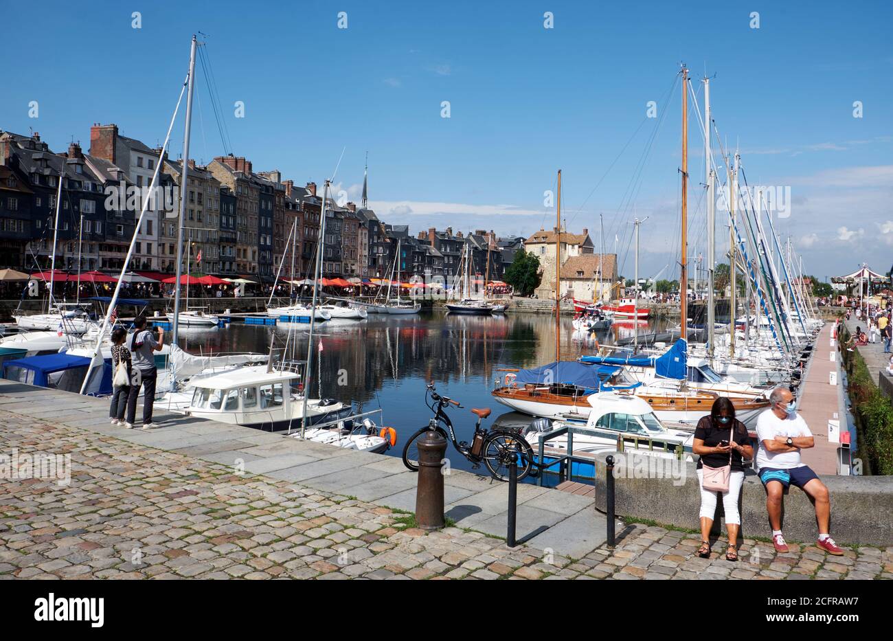 Couple sitting on wall wearing face masks during the COVID-19 pandemic in the picturesque harbour town of Honfleur in Normandy Stock Photo