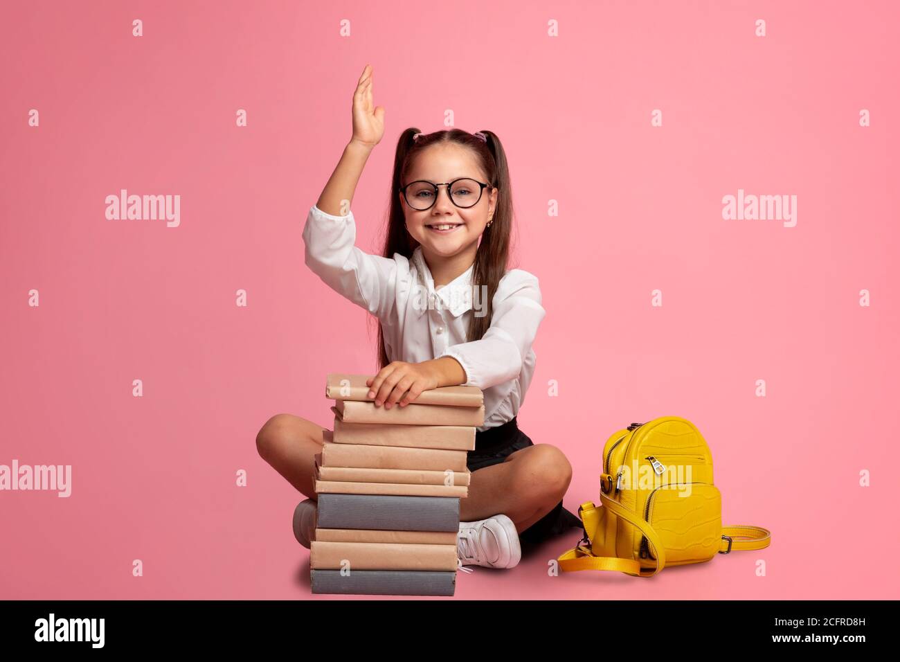 Clever pupil. Smiling girl in glasses with stack of books and backpack knows answer and raises hand up Stock Photo