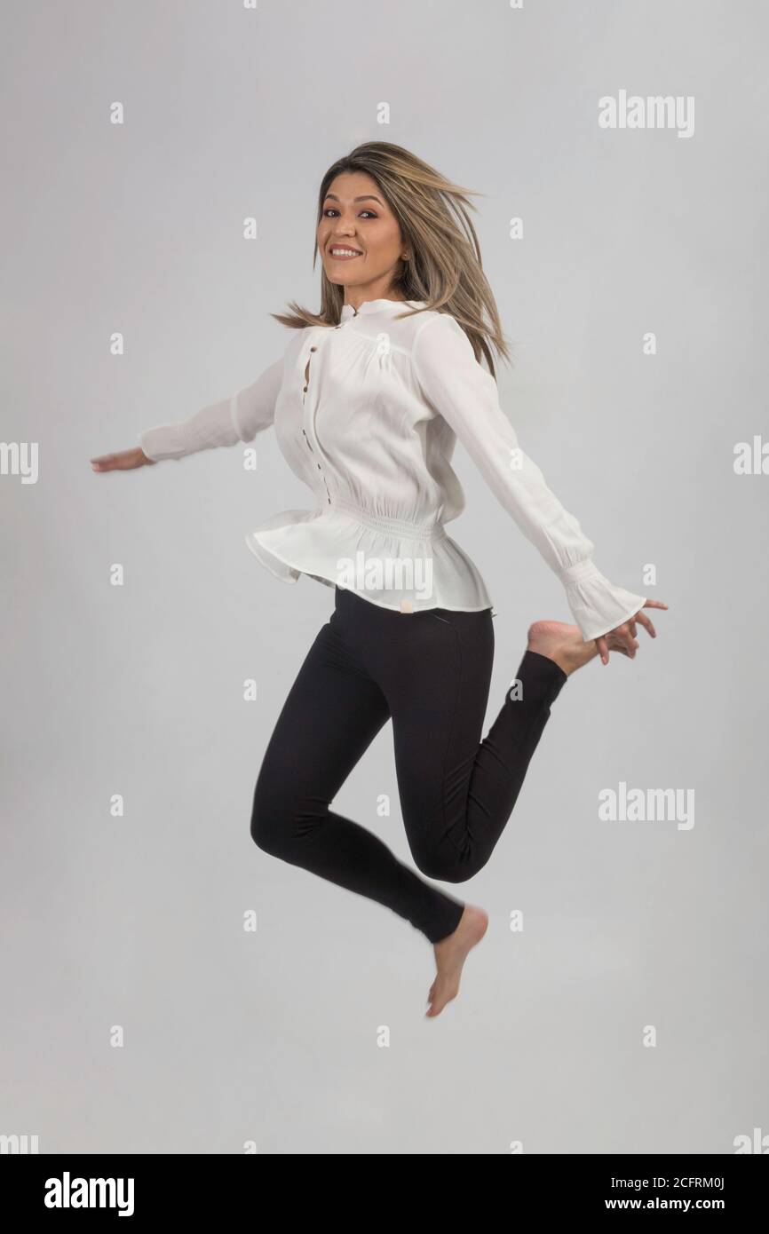 jumping and smiling young woman in white whithe blouse, black pants and barefoot, studio with gray background Stock Photo