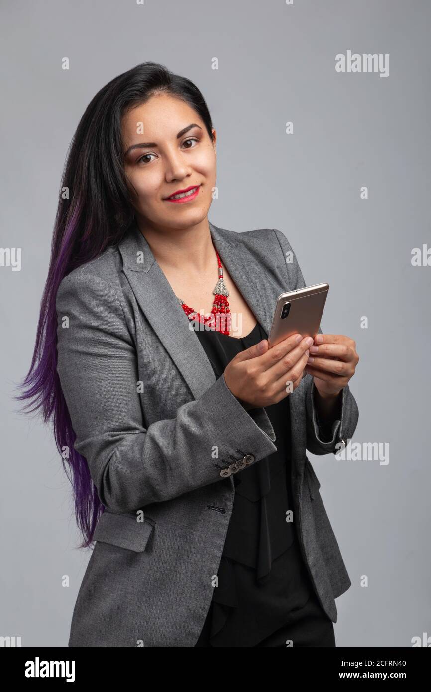 Latin young woman with long black hair using her cell phone, wearing gray formal jacket and red collar, studio with gray background Stock Photo
