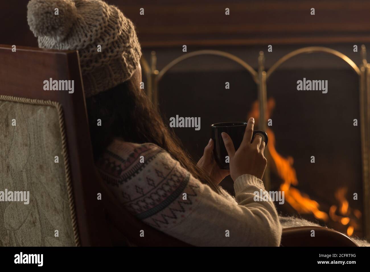 young Latin woman sitting on backwards, wearing a cap and wool sweater, in front of a lit fireplace, with a black cup in her hands Stock Photo