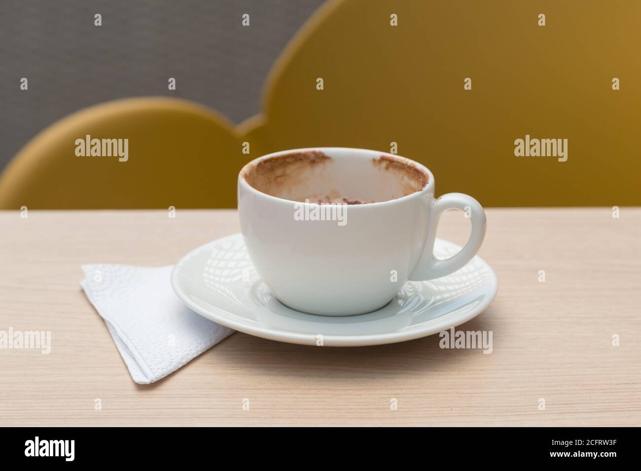 small white cup with chocolate, a plate and napkin, on a flat wooden surface, with a yellow chair in the background Stock Photo