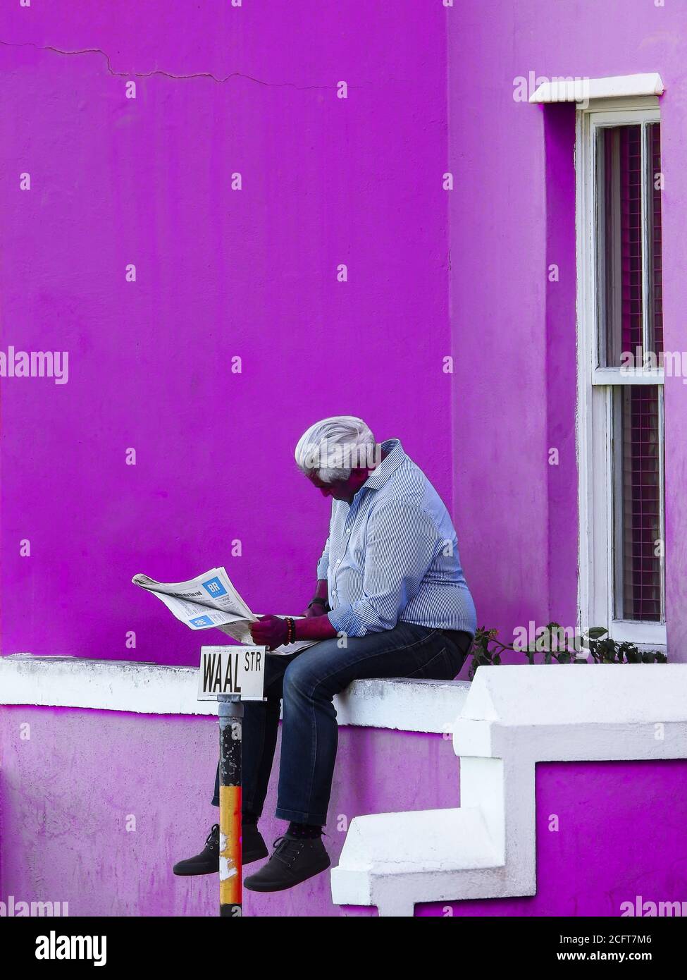 The daily life of a man: reading the newspaper sitting on the flower box of his purple house Stock Photo