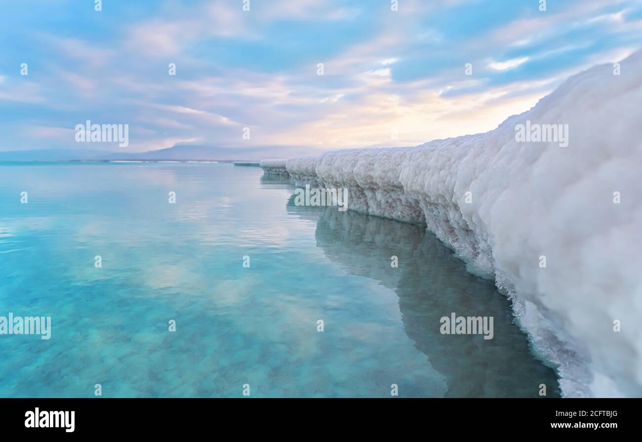 Sand completely covered with crystalline salt looks like ice or snow on shore of Dead Sea, turquoise blue water near, sky colored with morning sun Stock Photo