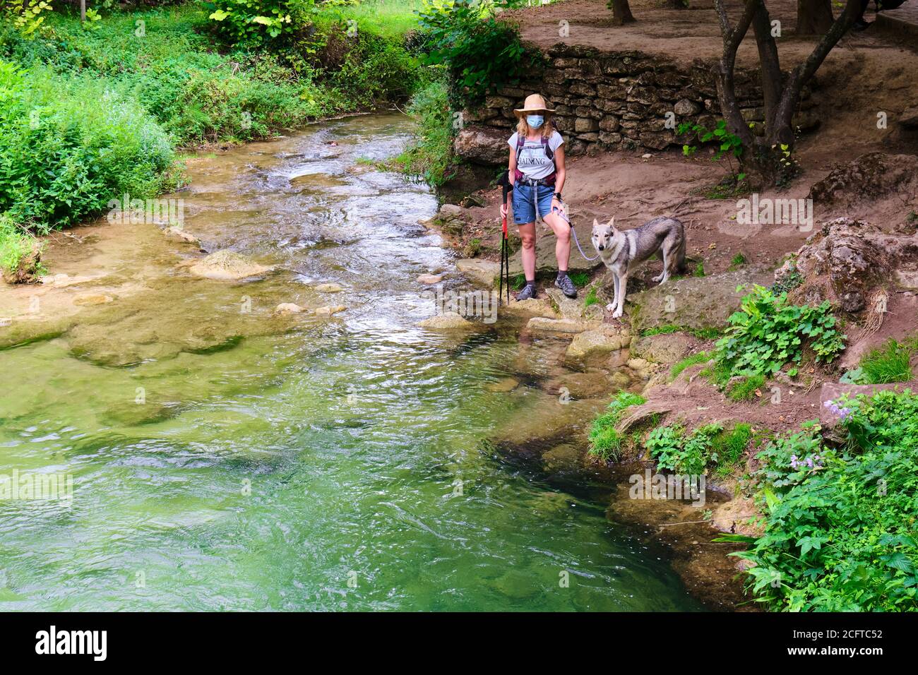 Hiker mature young woman with a face mask and a dog in a river bank. Stock Photo