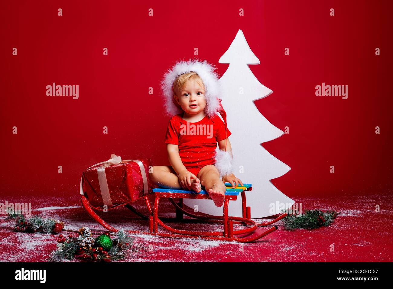 A little boy in a red Santa Claus T-shirt sits near a white Christmas tree with gifts Stock Photo