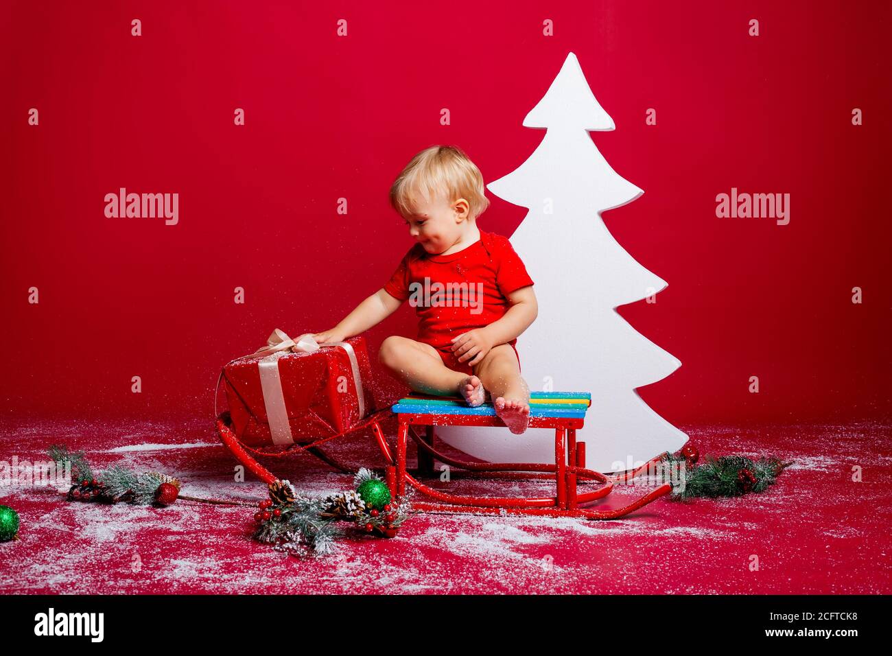 A little boy in a red Santa Claus T-shirt sits near a white Christmas tree with gifts Stock Photo