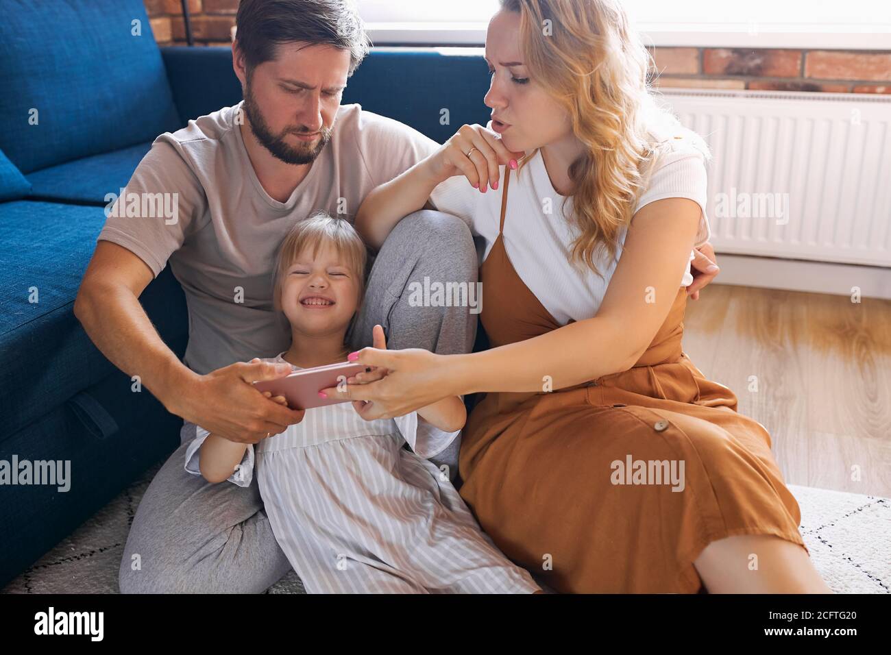 happy family with daughter watch funny video on mobile phone, girl is laughing, sit on the floor in living room Stock Photo