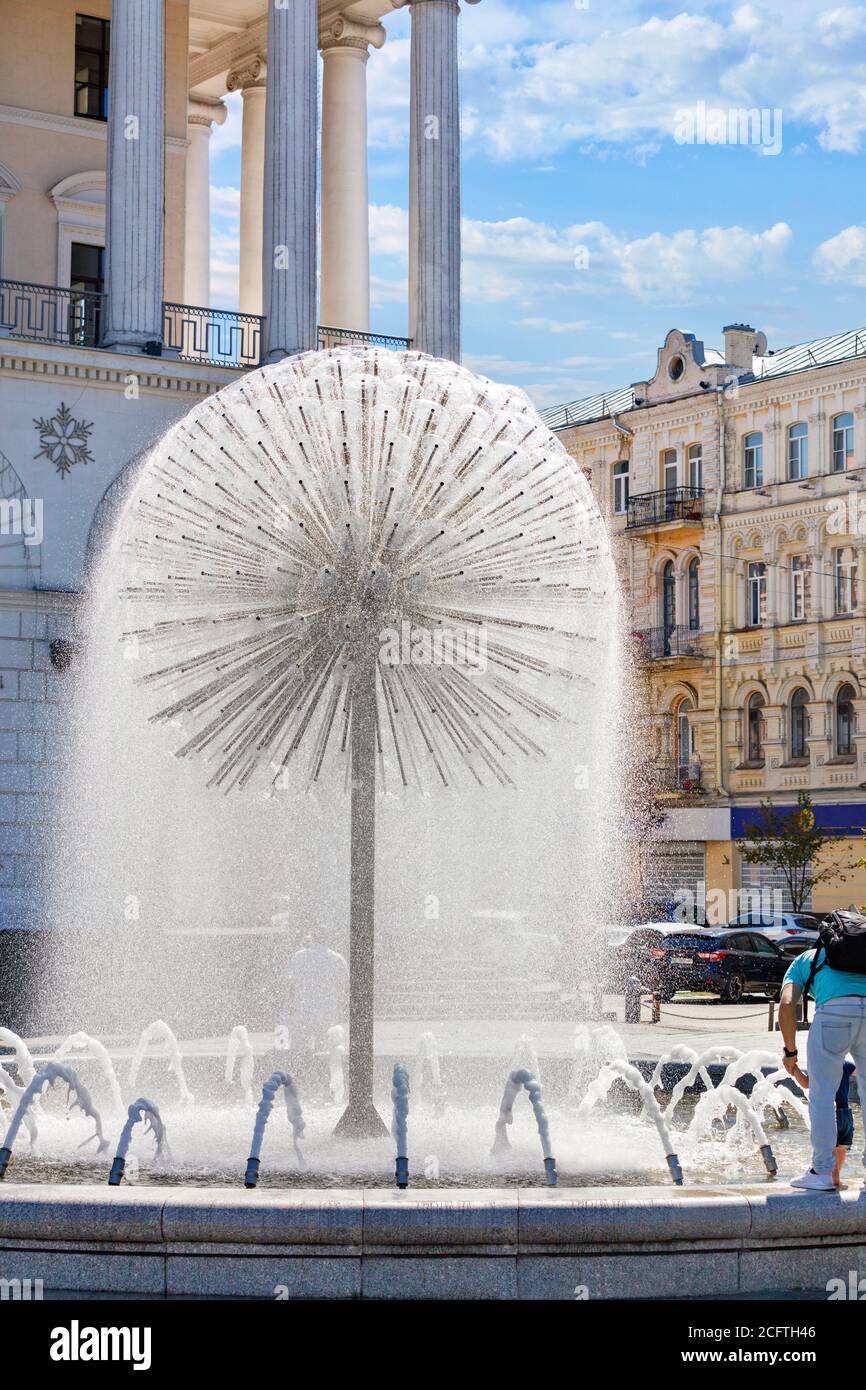 A light refreshing drizzling breeze from the city fountain in the shape of a huge dandelion gives people in the summer heat the freshness of water coo Stock Photo