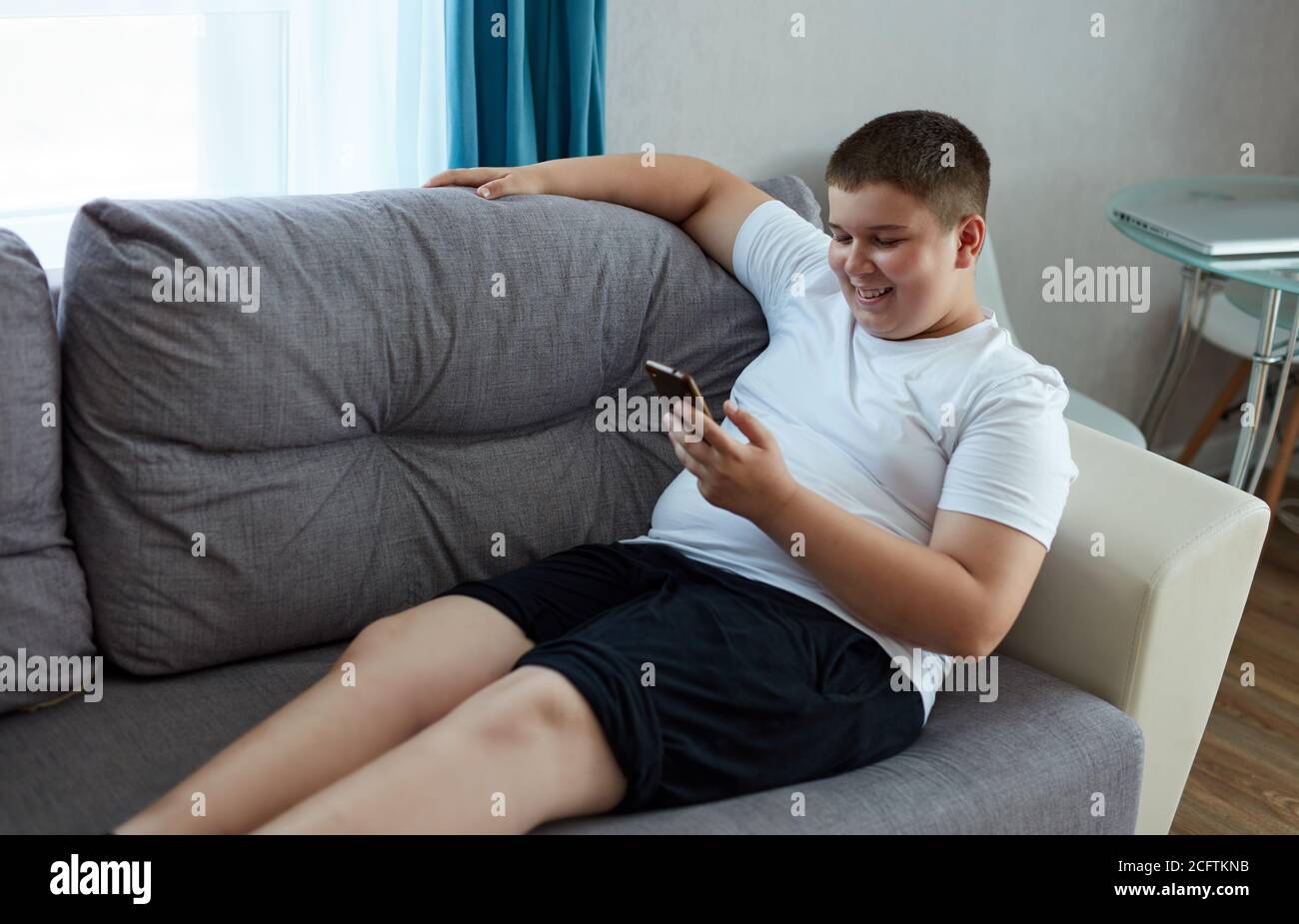 young fat boy chatting with friend, sit with mobile phone, teen boy in domestic wear sits on sofa and look at smartphone, smile. leisure time Stock Photo