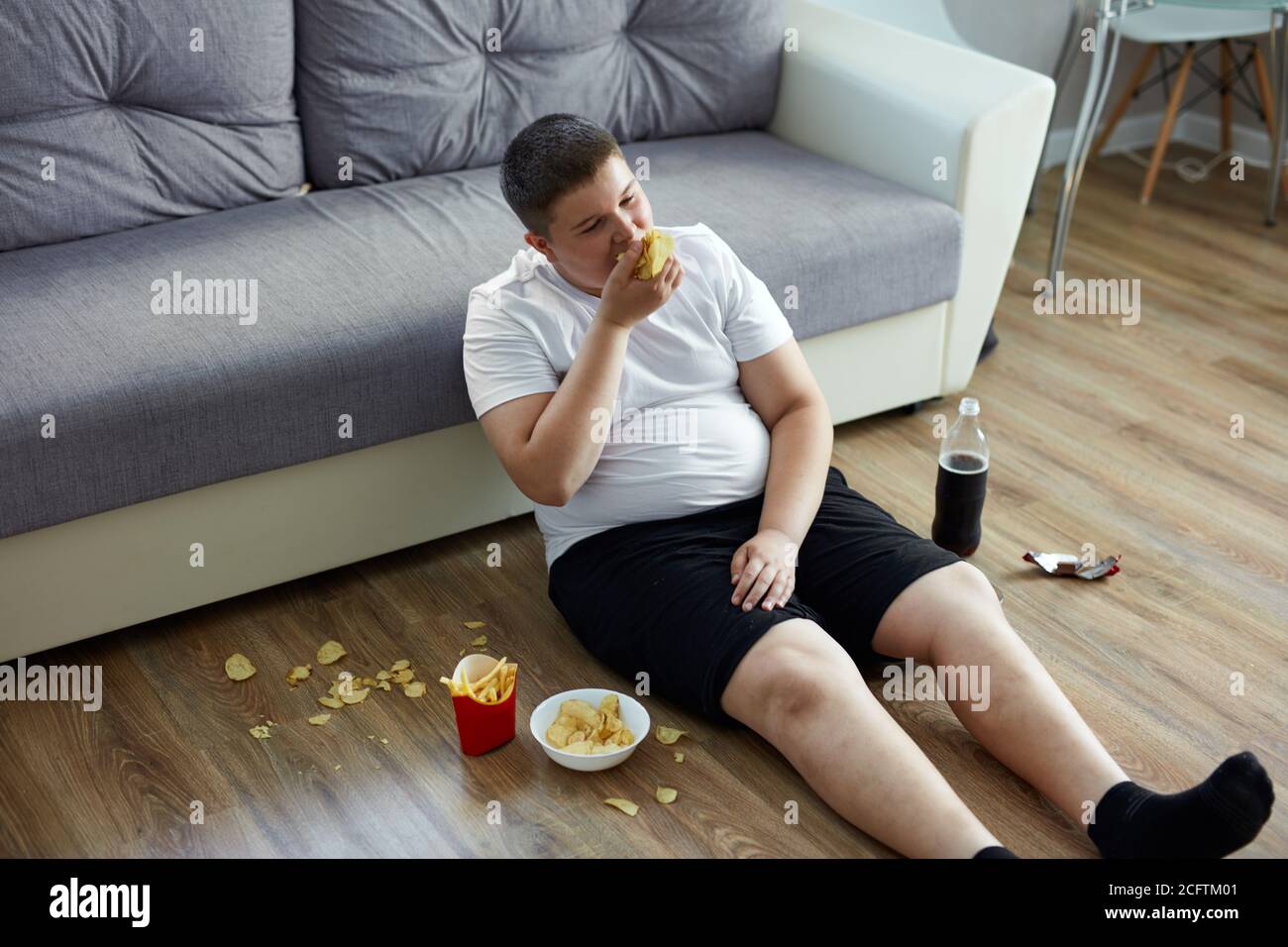 fat overweight teenager boy has bad nutrition, eat unhealthy food. sit on the floor eating junk food and watching tv Stock Photo
