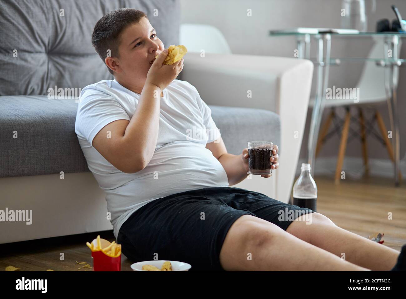 fat overweight teenager boy has bad nutrition, eat unhealthy food. sit on the floor eating junk food and watching tv Stock Photo