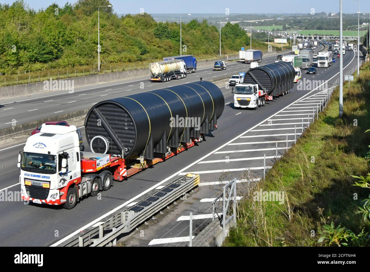 Wide oversize long cylinder shape load on lorry truck low loader occupying two lanes of M25 motorway with escort van at back with traffic queue UK Stock Photo