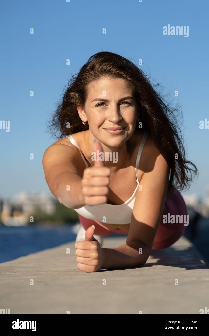 Fit joyful woman in pink legging doing plank exercise working on abdominal muscles and triceps, showing thumb up, outdoors. Sport female model doing c Stock Photo