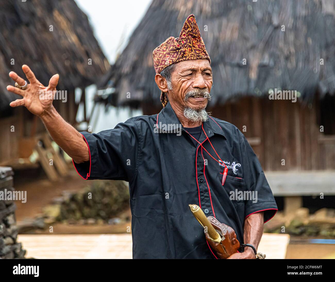 The village chief gives a welcome speech in a pointed ikat headdress, gesturing with a raised hand, a knife in a sheath tucked into his belt, his shirt bearing the lettering "Wonderful Indonesia", mountain village near Moni, Flores, Indonesia. Stock Photo