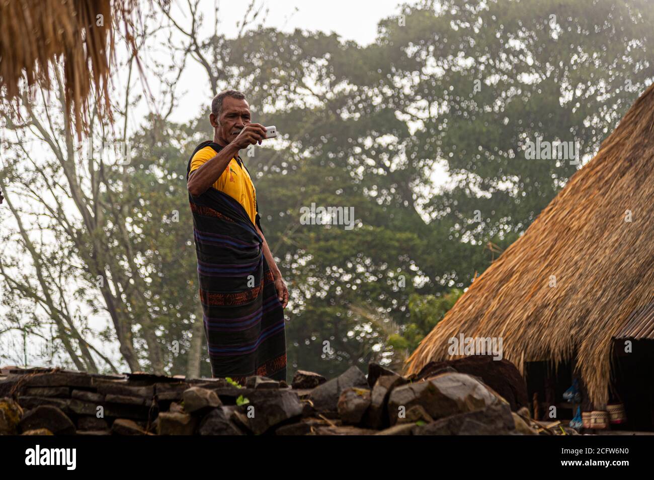 Amid a stack of stones and tall trees, a man in a yellow polo shirt over his ikat sarong takes a photo with his smartphone in a mountain village near Moni, Flores, Indonesia. Stock Photo