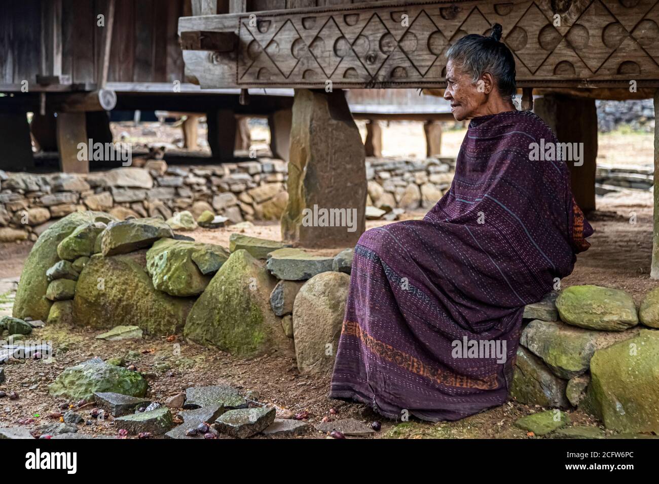 A woman in purple ikat clothing sits on a stone wall beneath a lumbung storage house raised on stilts in the village of Wologai, Flores, Indonesia. Such stilt buildings traditionally serve for storing provisions. Stock Photo