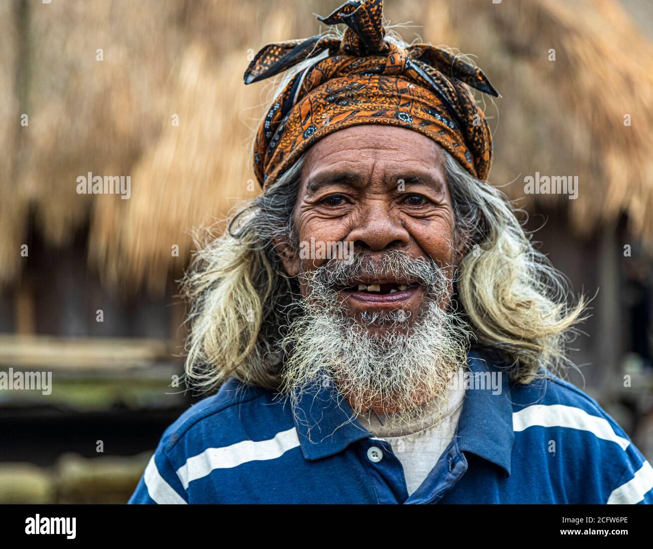 An old man with a grey beard and an orange batik headscarf smiles at the camera with gaps in his teeth, his long grey hair falling over the shoulders of a blue-and-white striped shirt, mountain village near Moni, Flores, Indonesia. Stock Photo