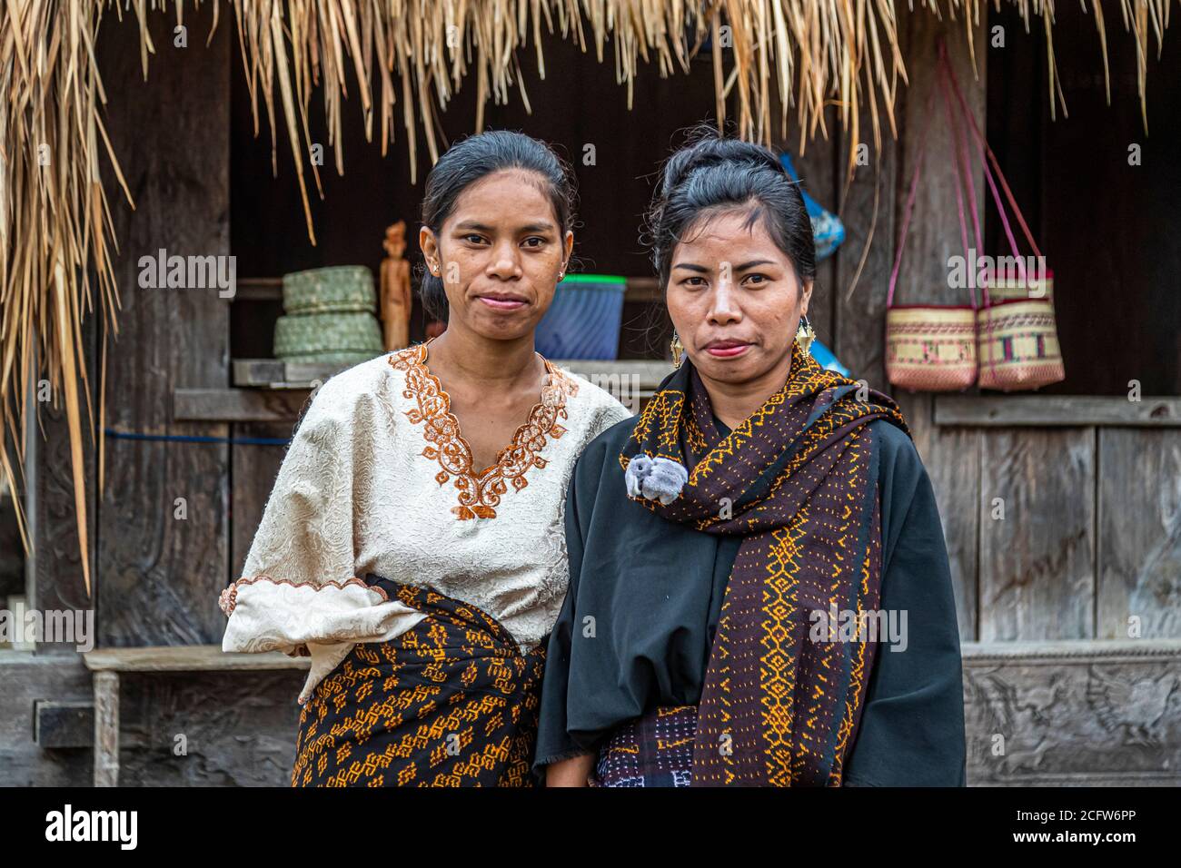 Two women in traditional ikat clothing pose in front of a wooden house with woven bags hanging on the wall in a mountain village near Moni, Flores, Indonesia. Stock Photo
