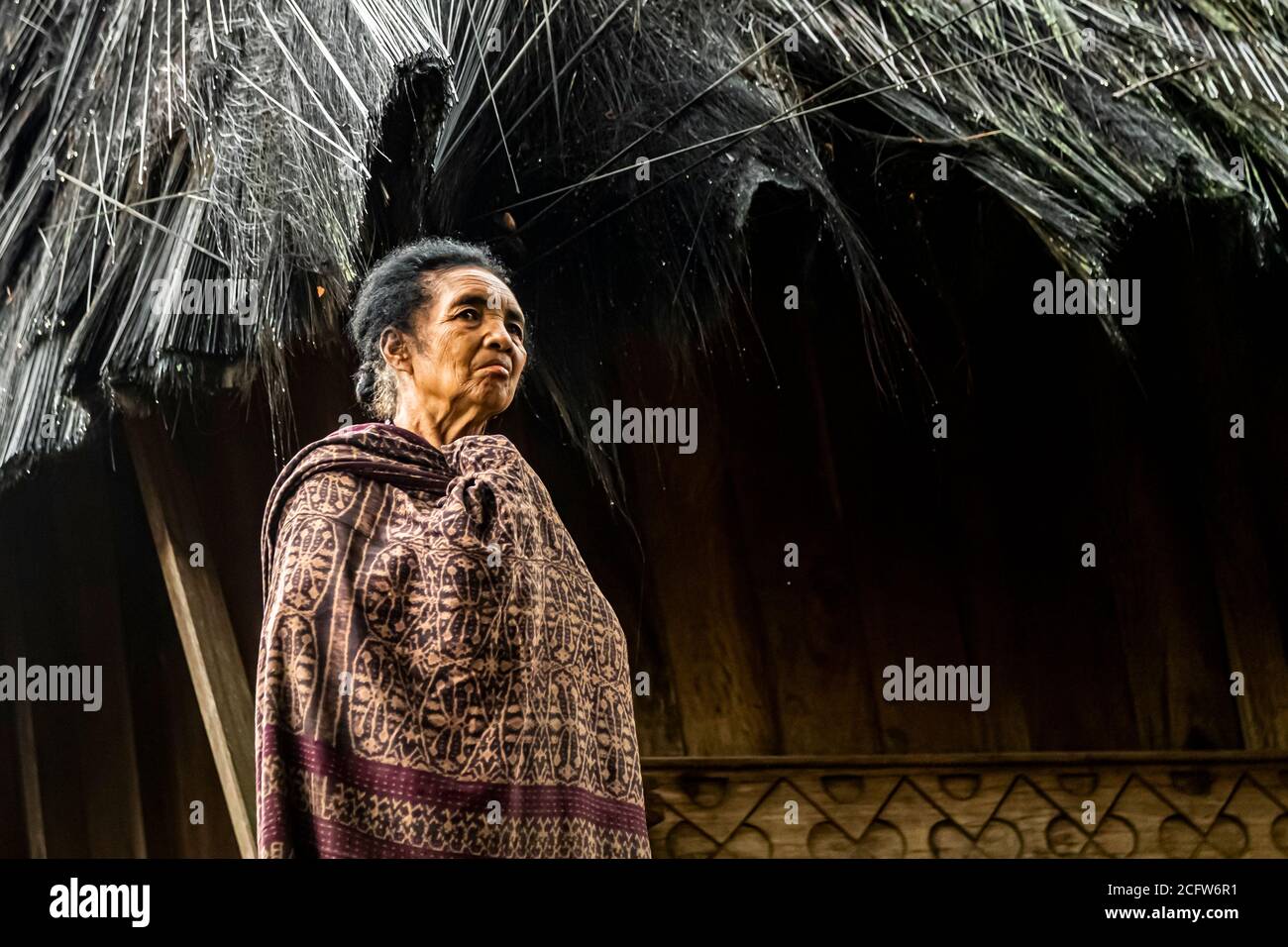 A woman in patterned ikat dress stands beneath the thatched roof of a house in the village of Wologai, Flores, Indonesia, looking to the side. The Lio village ranks among the oldest in the Ende region. Stock Photo