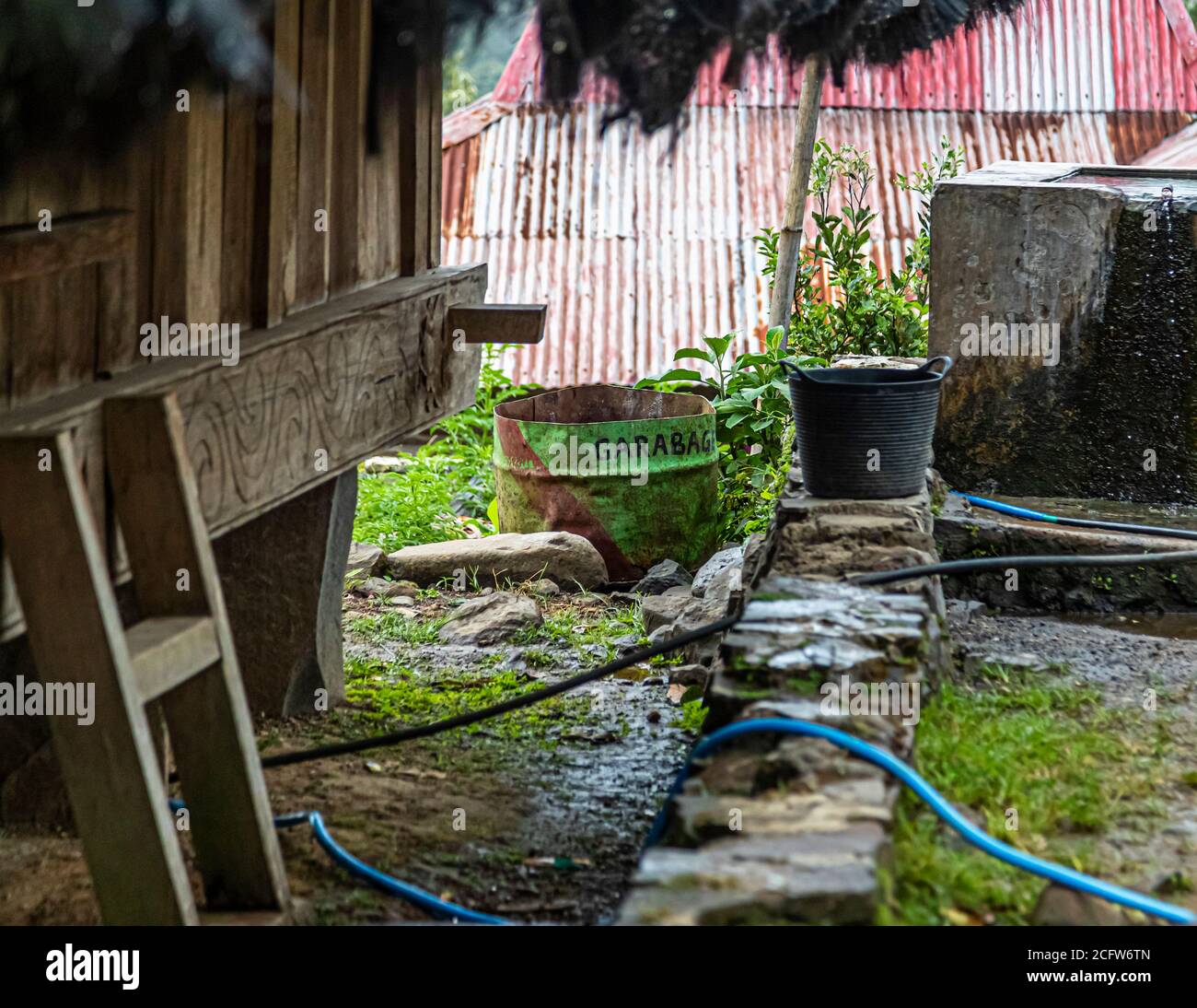 A green-painted barrel with the hand-lettered word "Garabag" (garbage) serves as a rubbish bin in the village of Wologai, Flores, Indonesia. Water hoses and a stone-lined water channel run through the yard in front of it. Stock Photo