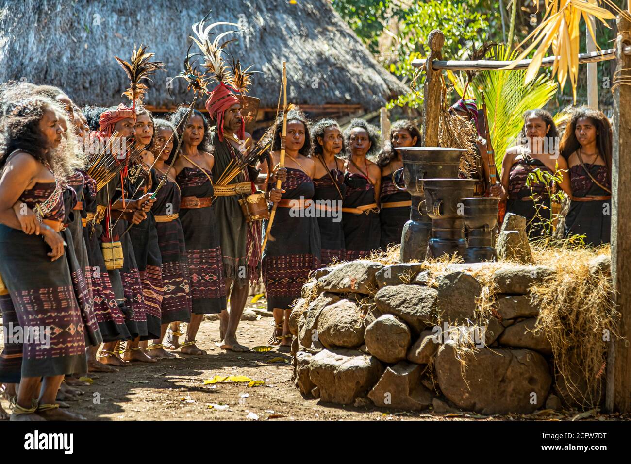 The feather-crowned leader stands with bow and arrow in front of the bronze Moko drums as the dancers wait in a line behind him, in the village of Takpala, Alor, Indonesia. Stock Photo