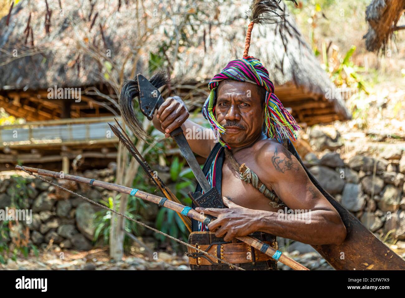 With his machete raised and a piercing gaze, the tattooed warrior poses in front of a thatched roof, village of Takpala, Alor, Indonesia. Stock Photo