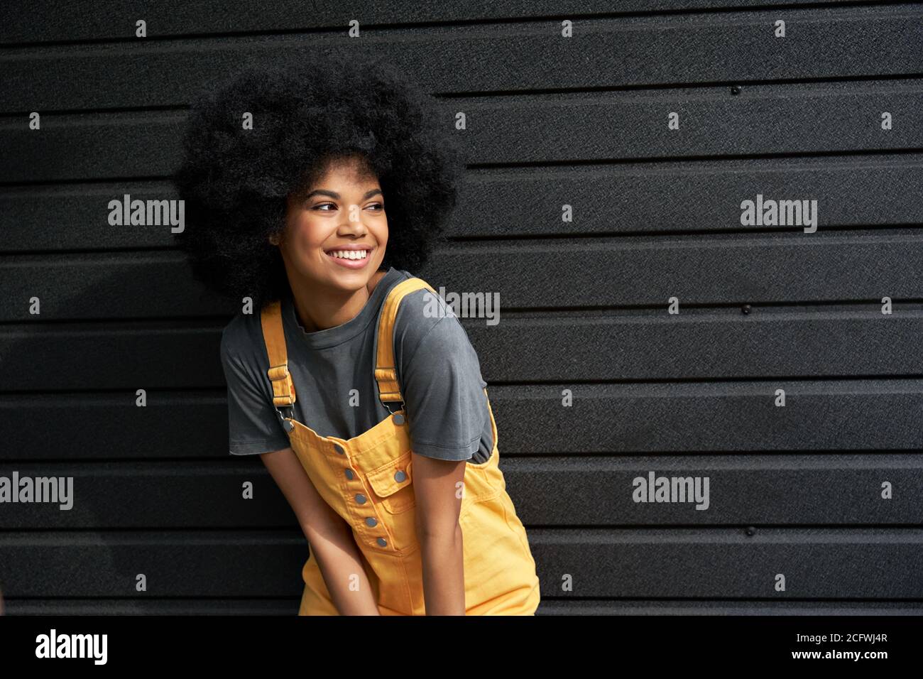 Happy stylish African lady with afro hair looking away stand against black wall. Stock Photo