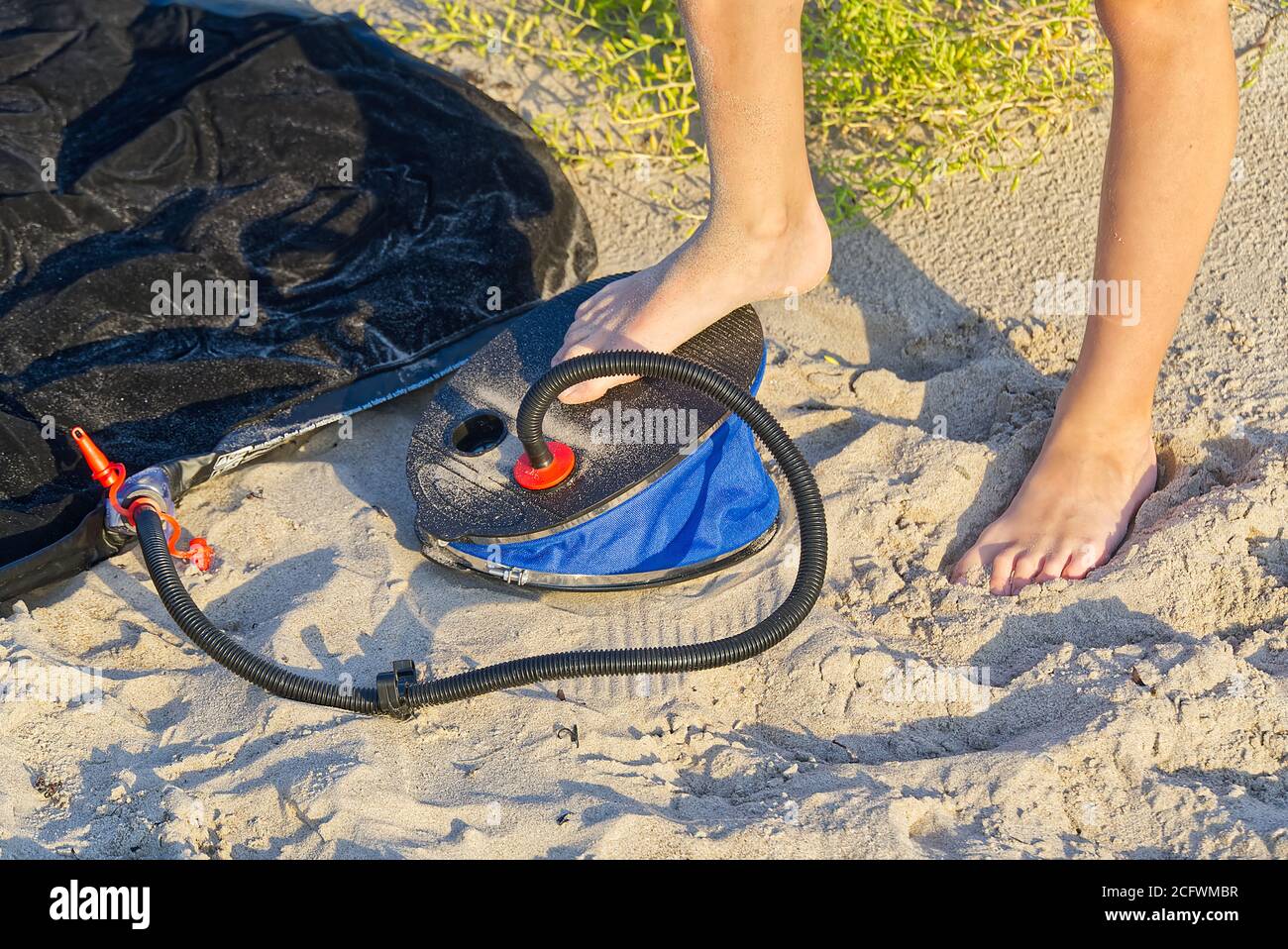 A Man with air foot pump pumps an inflatable mattress or air bed at sandy beach. Foot inflates air mattress with foot pump on sand. Stock Photo