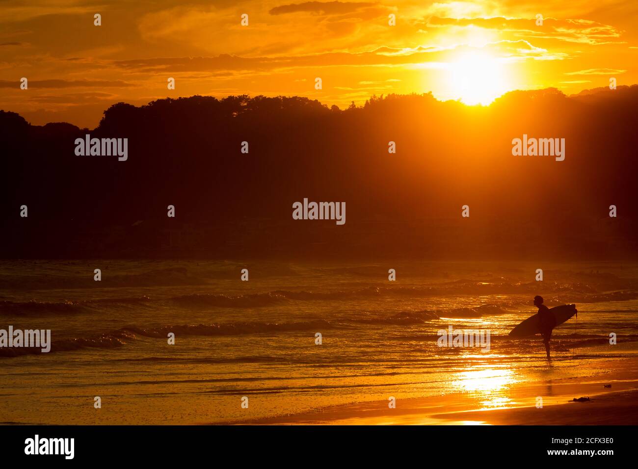 A surfer stands on a beach looking at the waves as the sun sets behind, Kamakura beach, Kanagawa, Japan. Stock Photo