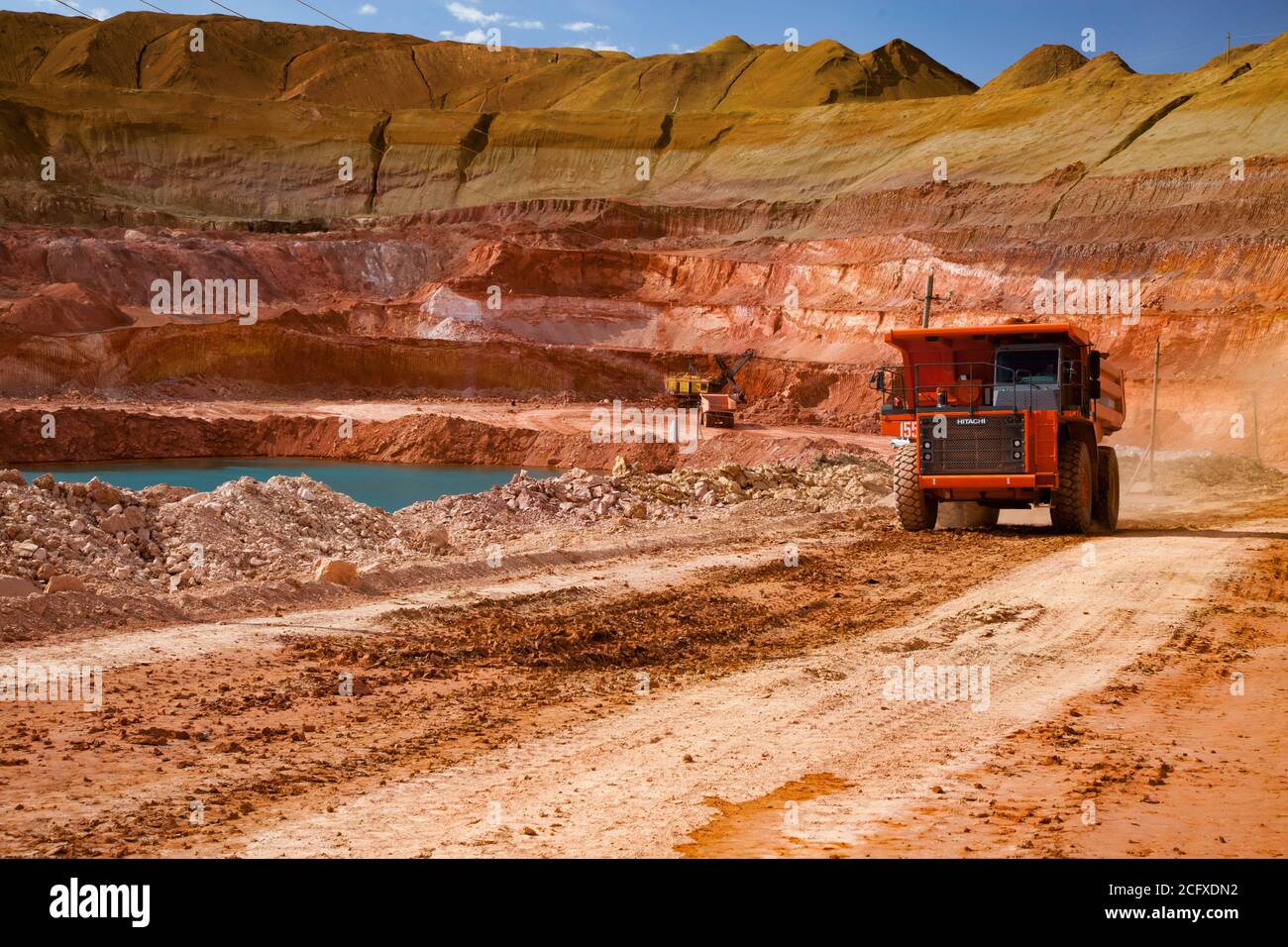 Aluminium ore mining and transporting. Open-cut quarry mining. Orange Hitachi dump-truck. Blue quarry lake. Blue sky with white light clouds. Stock Photo