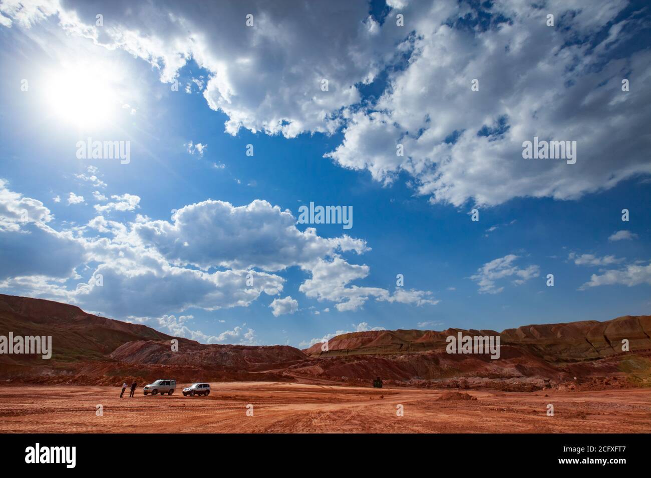 Red bauxite clay. Aluminium ore mining. Quarry of red ground on blue sky with clouds. Two off-road cars and two people. Stock Photo