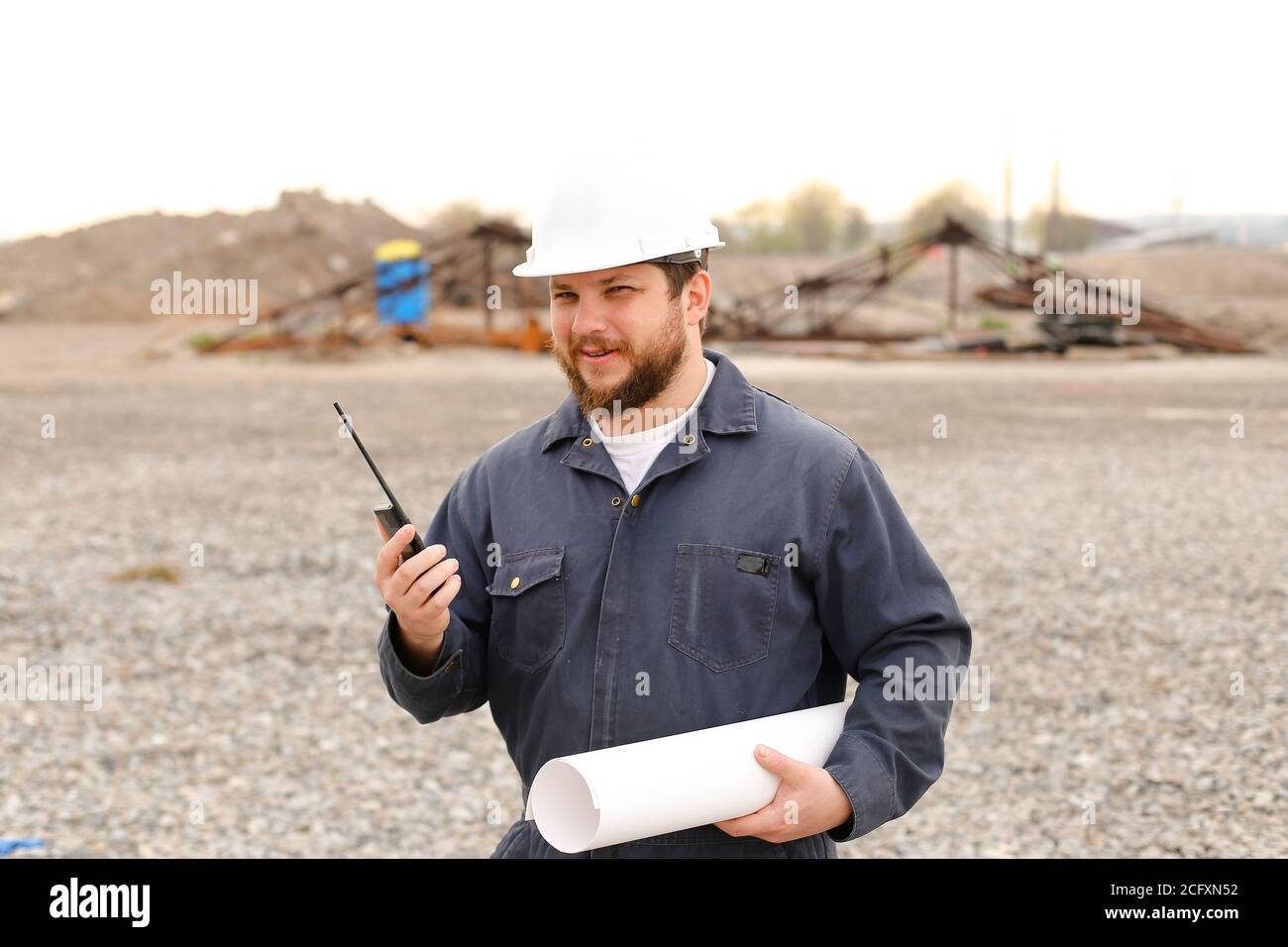 Portrait of caucasian foreman standing on construction site, talking by VHF walkie talkie and holding blueprints. Stock Photo