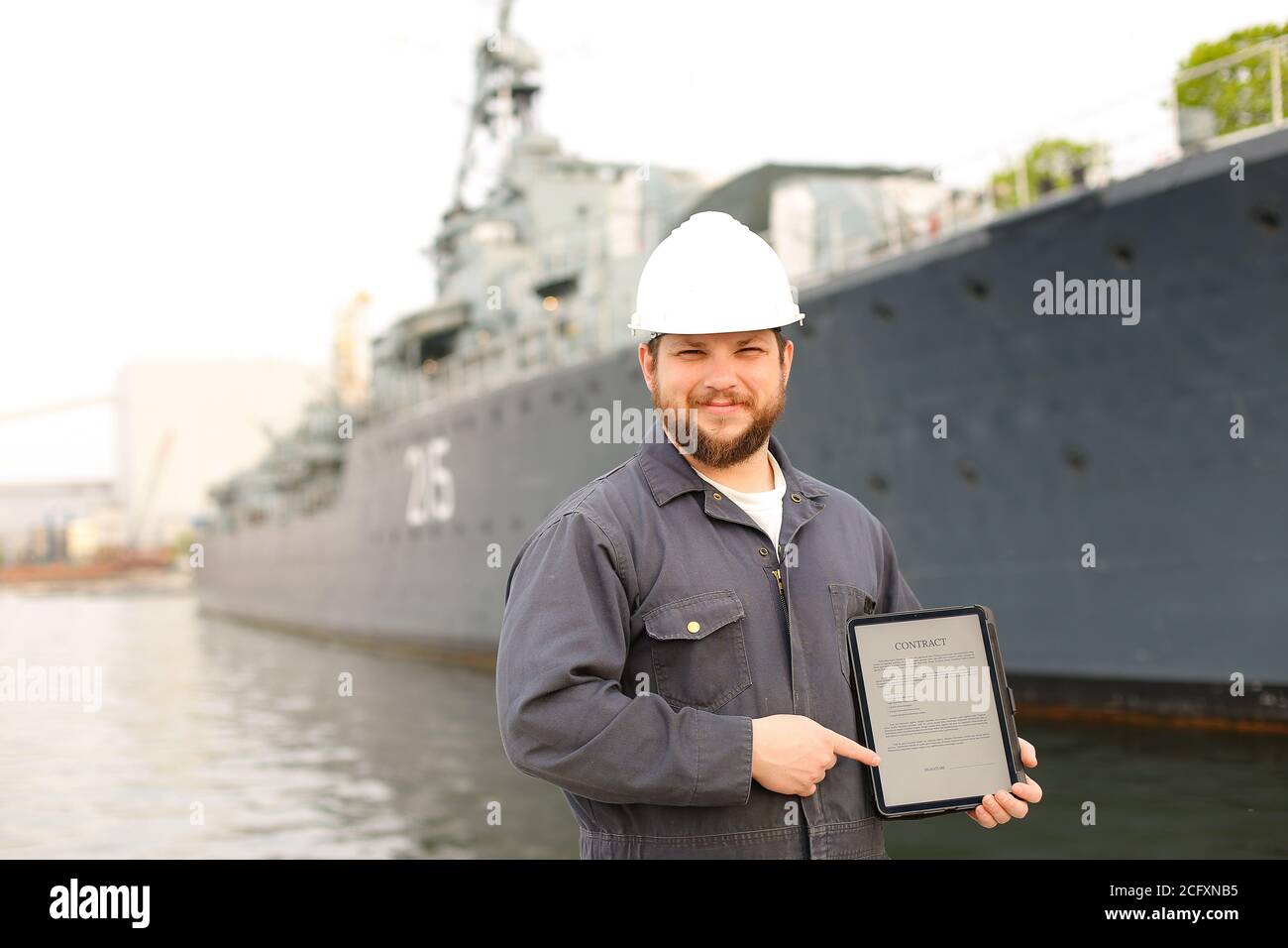 Captain in helmet showing contract on tablet near vessel in background. Stock Photo