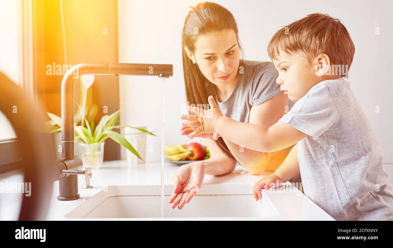 Child learning to wash hands with mother at the sink in the kitchen as hygiene precaution Stock Photo