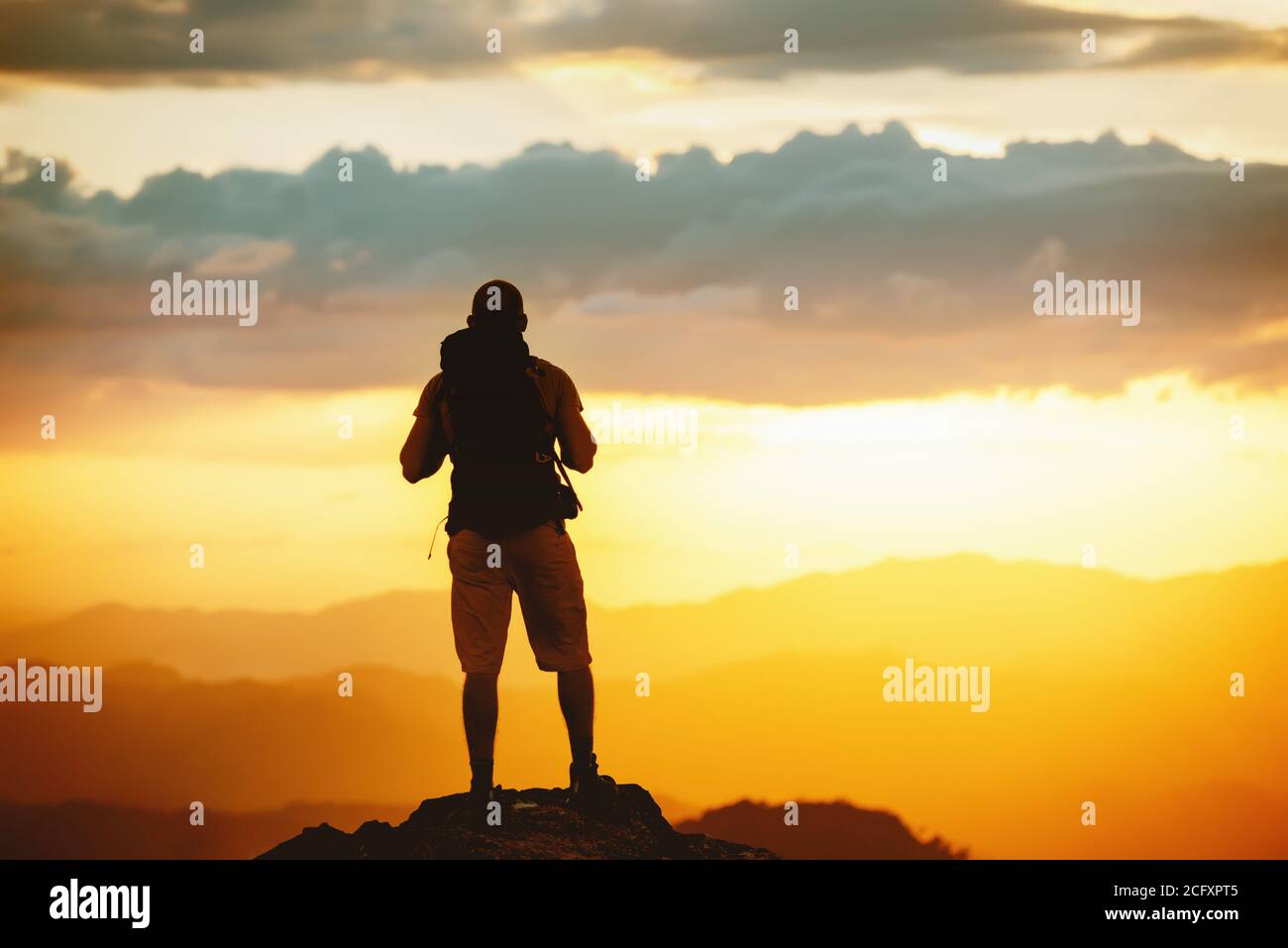 Unrecognized hiker stands with backpack on mountain top and enjoys sunset sky Stock Photo