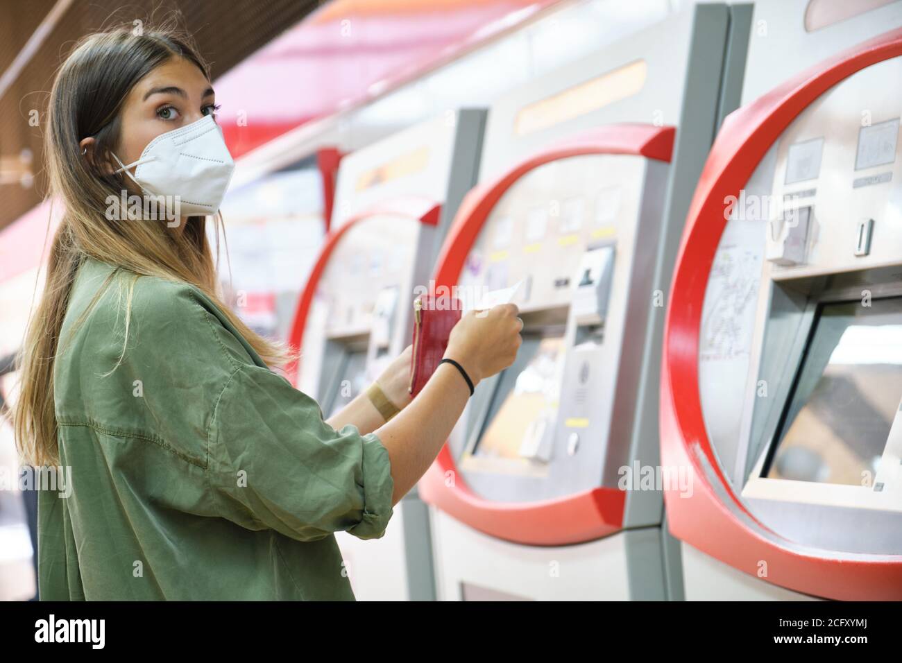 Young woman wearing face mask, buying a ticket at the train machines ...