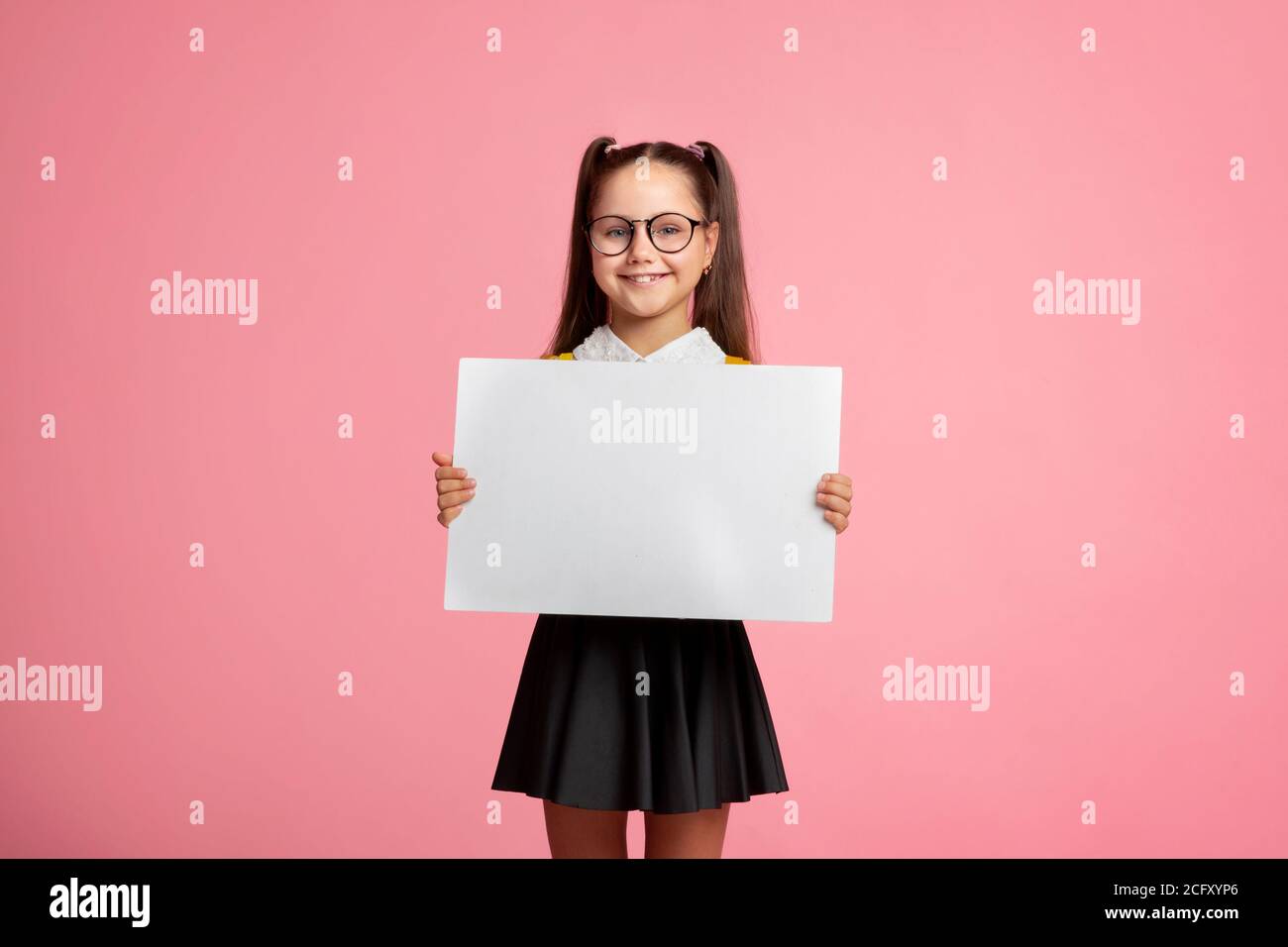 Cheerful child schoolgirl in uniform and glasses holds sign with empty space Stock Photo