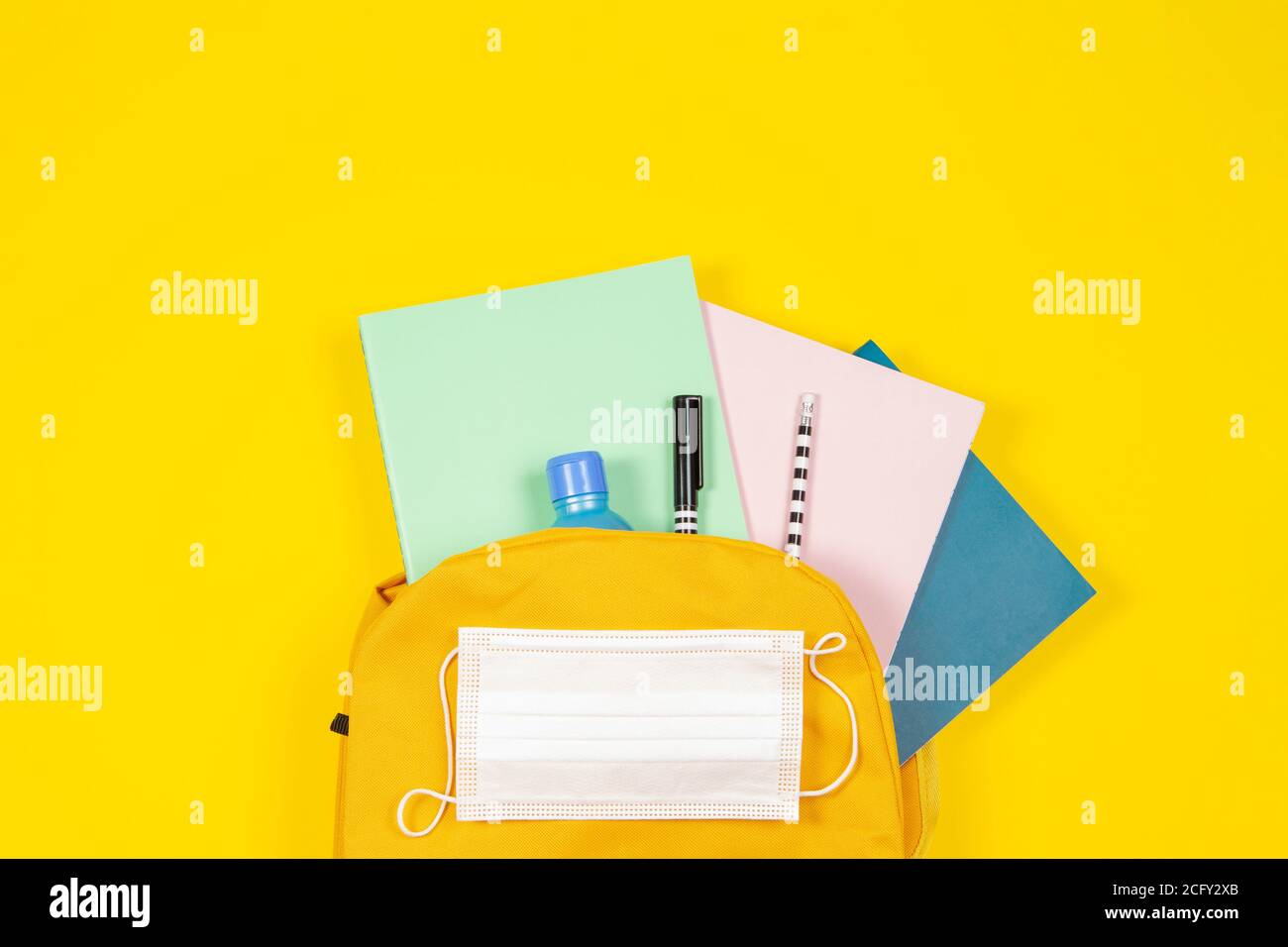 Education, learning, back to school during pandemic. Top view to backpack with school supplies, hand sanitizer, protective mask on yellow background Stock Photo