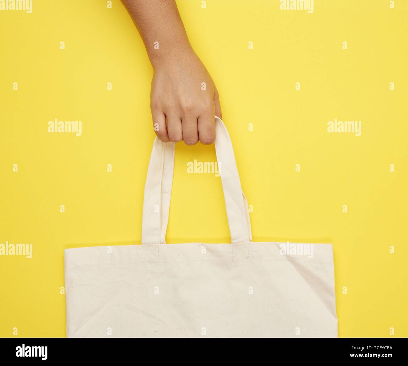 Female Hand Holds An Empty Beige Textile Bag By The Handles On A Yellow female-hand-holds-an-empty-beige-textile-bag-by-the-handles-on-a-yellow
