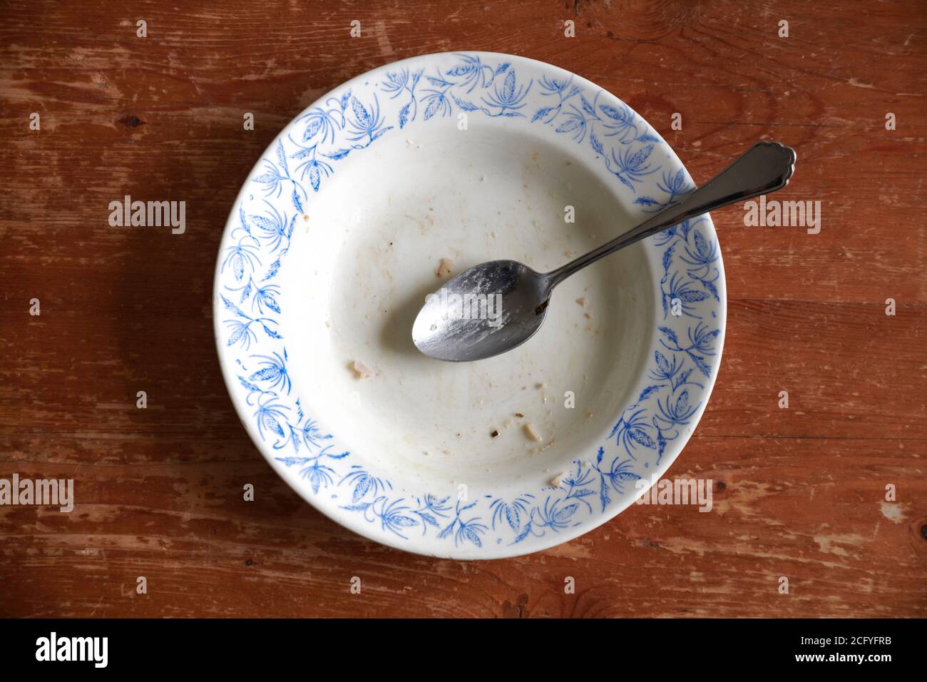 Ein leer gegessener weisser, tiefer Teller mit blauen, floralem Muster mit einem silbernen Loeffel steht auf einer braunen, stark beanspruchten Tischp Stock Photo