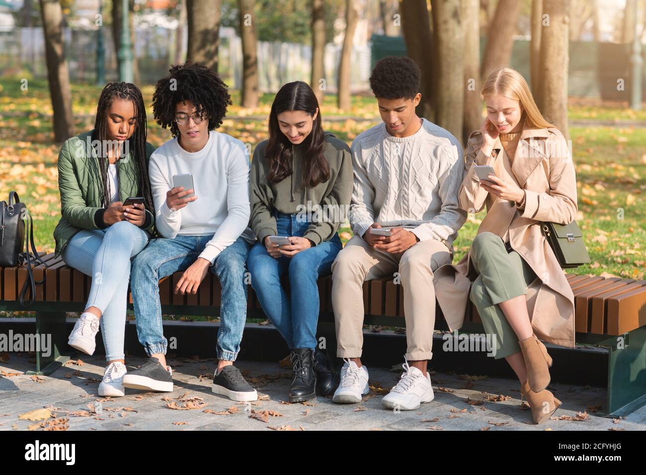 Teenagers sitting on bench in park and using smartphones Stock Photo