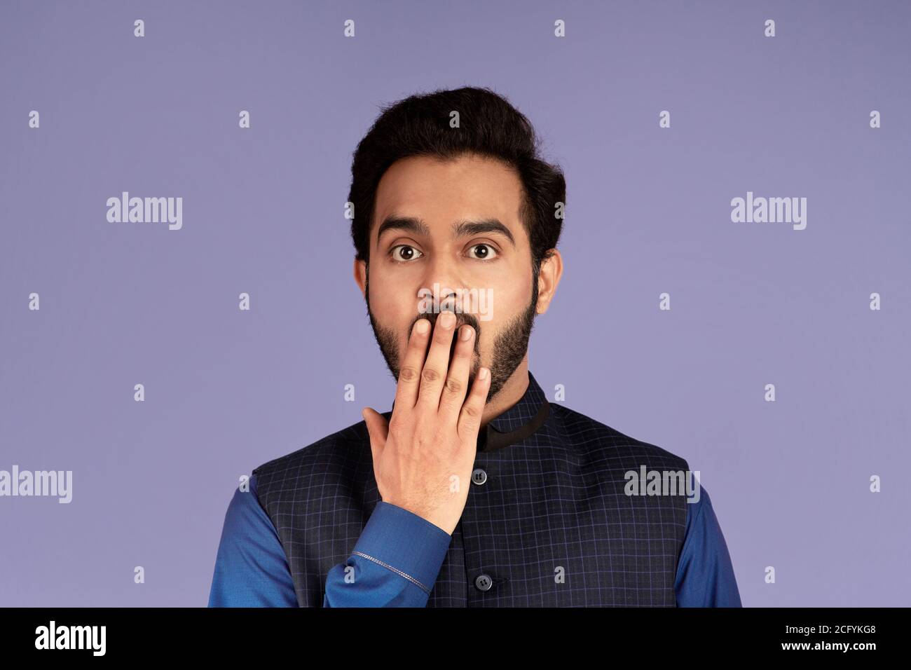 Silence or mistake concept. Shocked Indian man covering mouth with his hand over lilac background Stock Photo