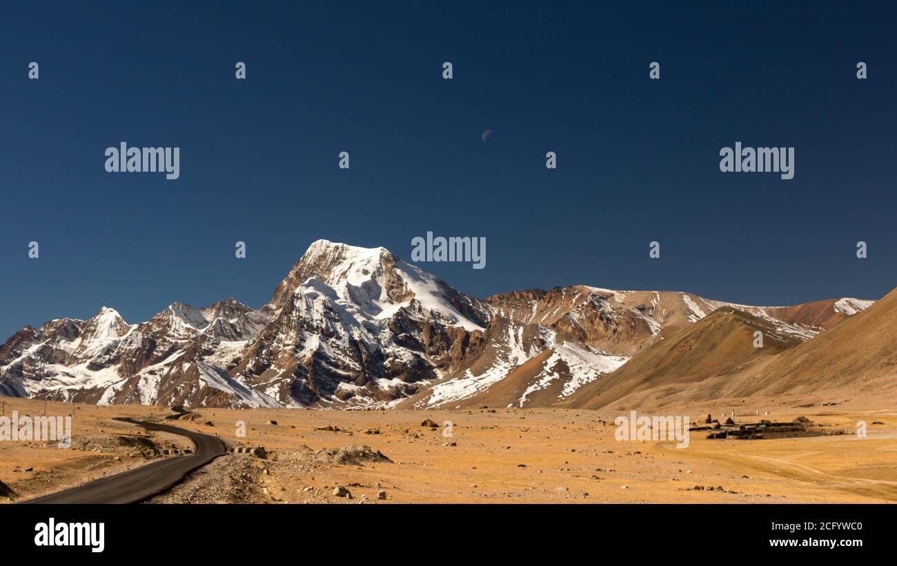 Selective focus image of a long lonely road with curves on the tibetan plateau with snow clad mountains and peaks and clear blue sky in front and tibe Stock Photo