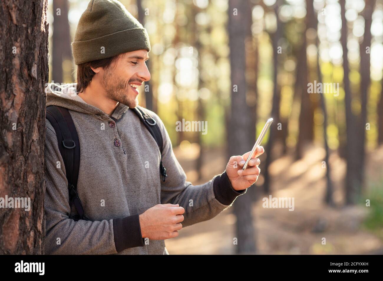 Positive guy leaning on tree in forest, using mobile phone Stock Photo